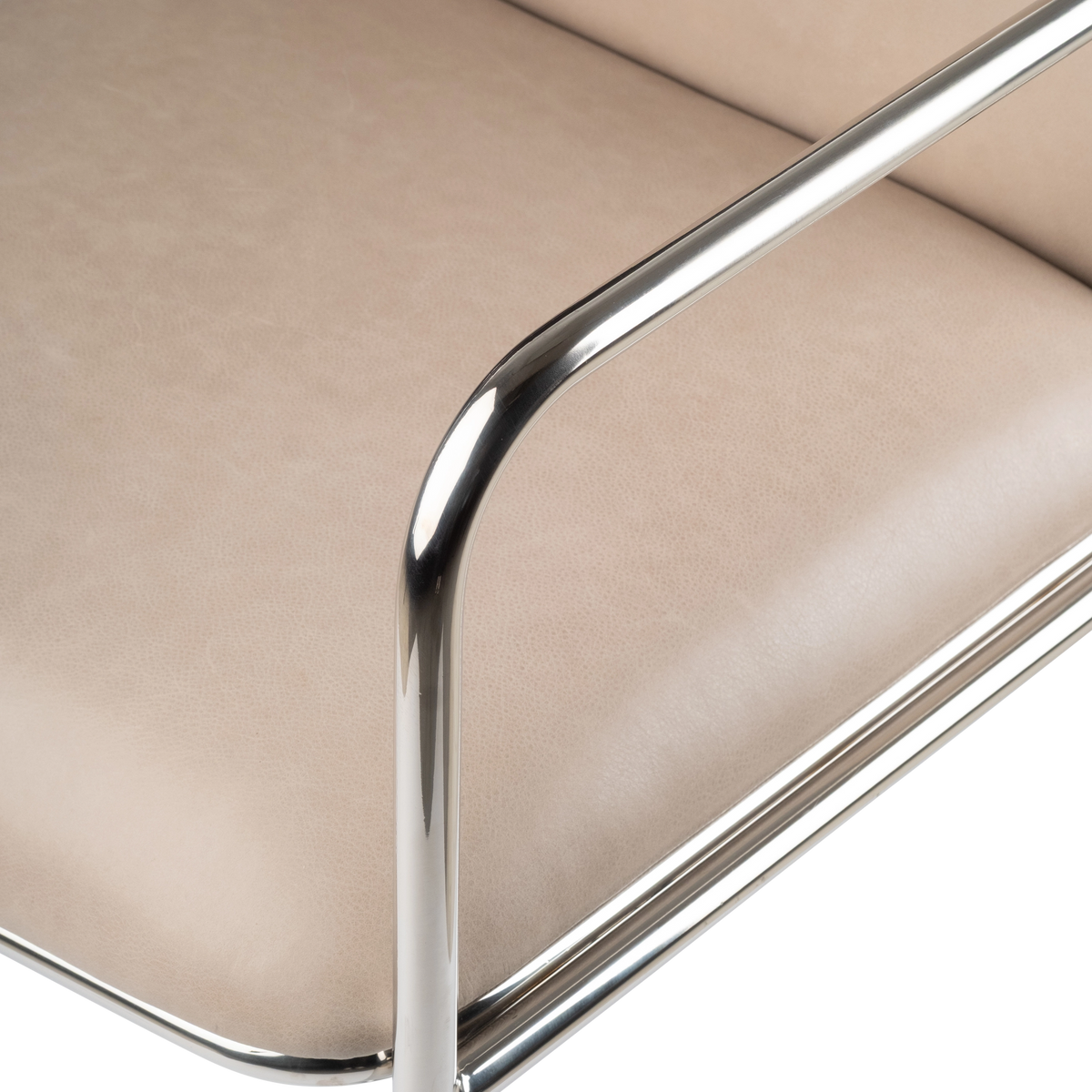 Close-up of the Rhodes Arm Chair’s polished stainless steel arm curving over the taupe leather seat, with the tubular frame reflecting soft highlights.