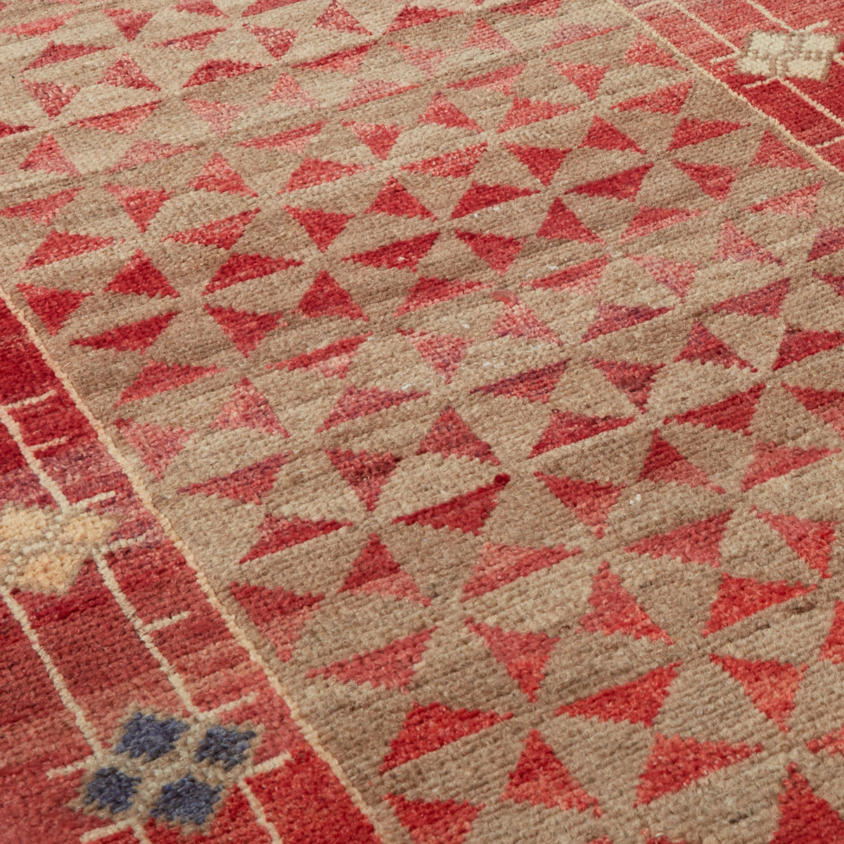 Close-up of a woven rug with a geometric pattern of red triangles on a beige background, bordered by red stripes and small blue accents.