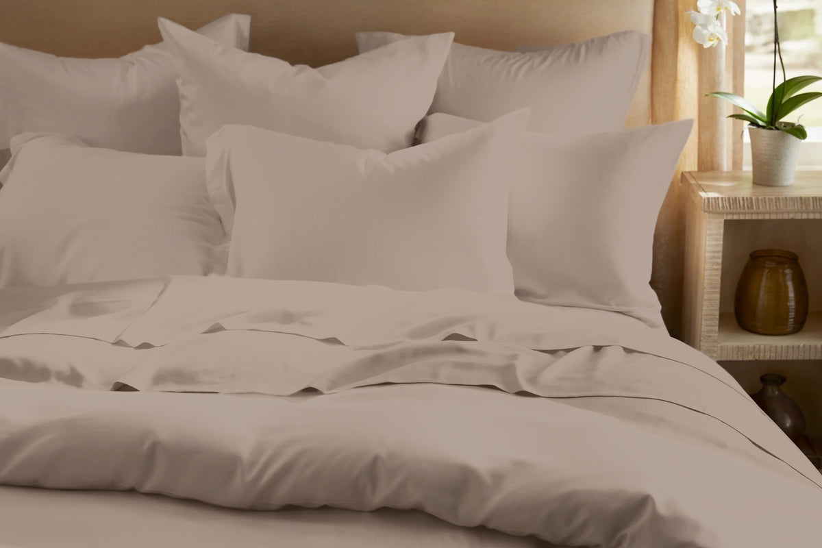 Front view of the Legna Classic Bedding Set in Mushroom, a muted taupe with a soft satin finish, showing euro and lumbar pillows, a folded-back flat sheet, and a smooth duvet cover against a warm wood headboard.
