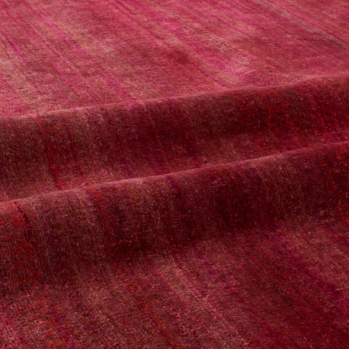 A close-up of a textured fabric or rug in deep burgundy and magenta tones, showing a soft woven surface with subtle horizontal striations and draped folds.
