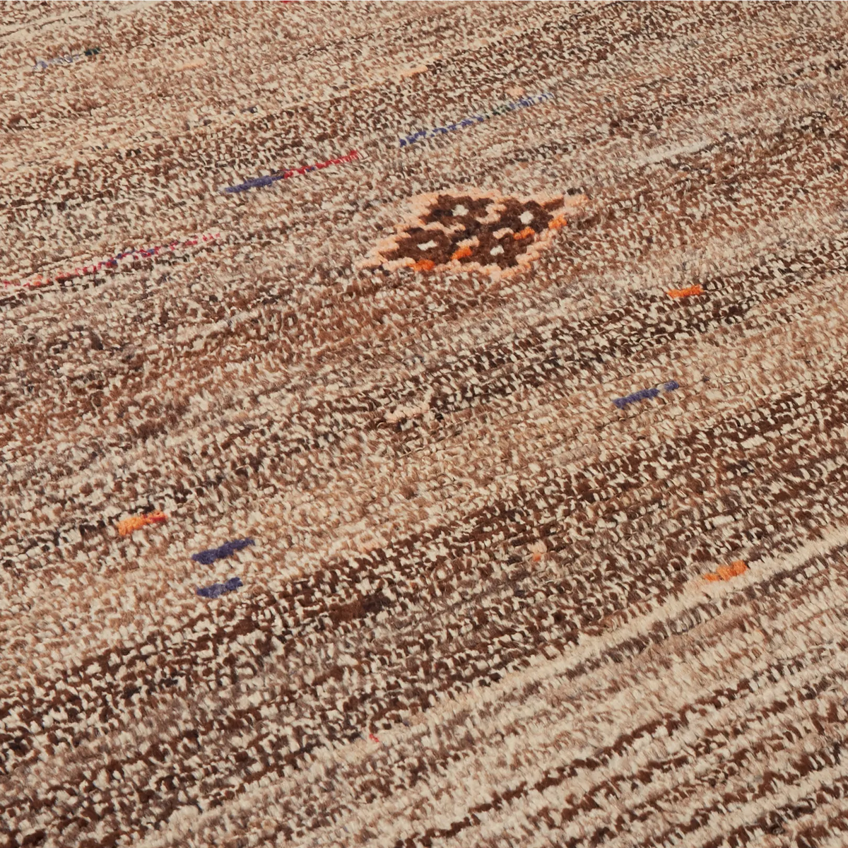 A close-up of a textured woven rug or carpet with horizontal striped patterns in brown, beige, cream, and small scattered accents of blue and orange.