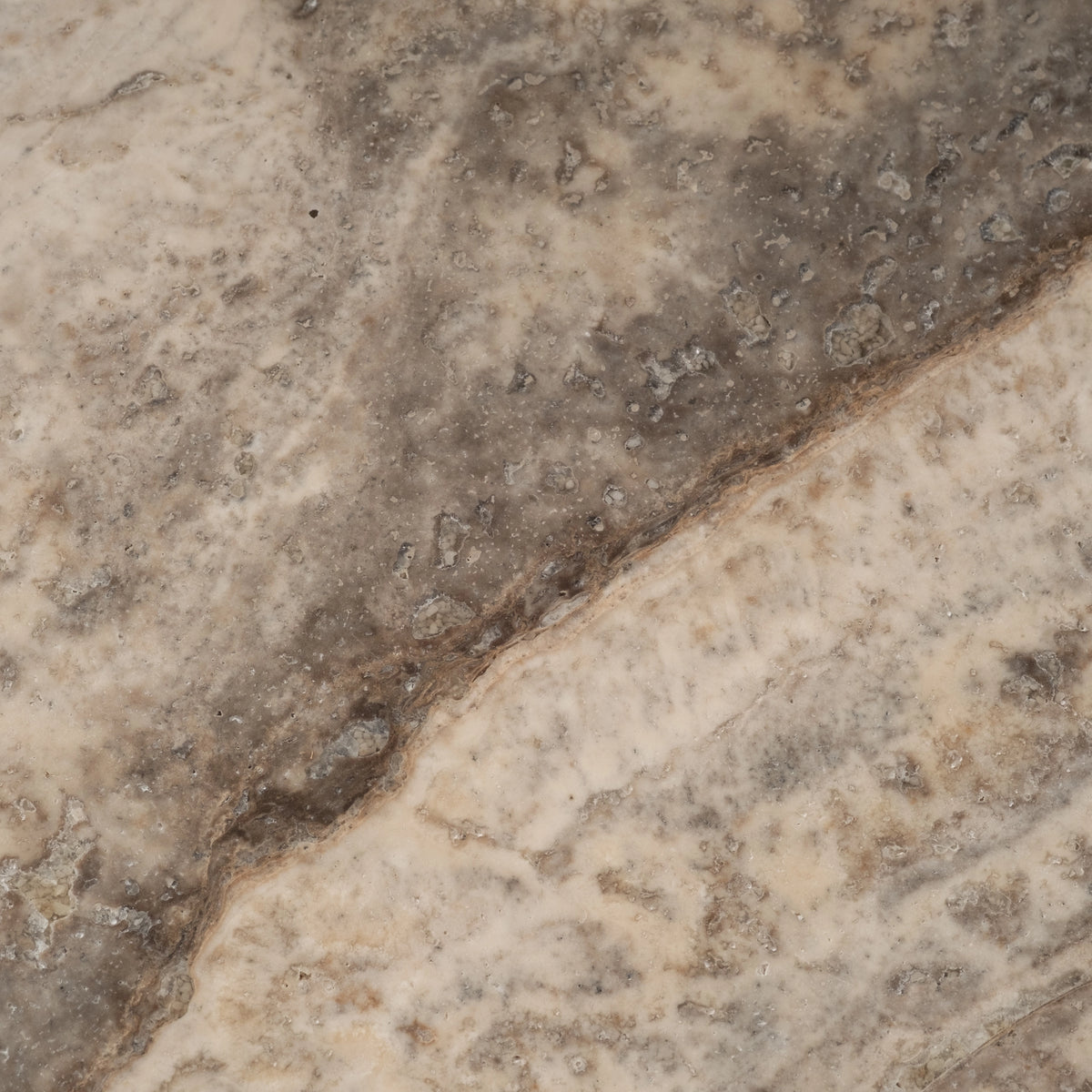 Close-up of the honed top of the Aldo Coffee Table in Taupe travertine, with a diagonal brown seam, small natural voids, and pale cream veining against a grey-taupe ground.