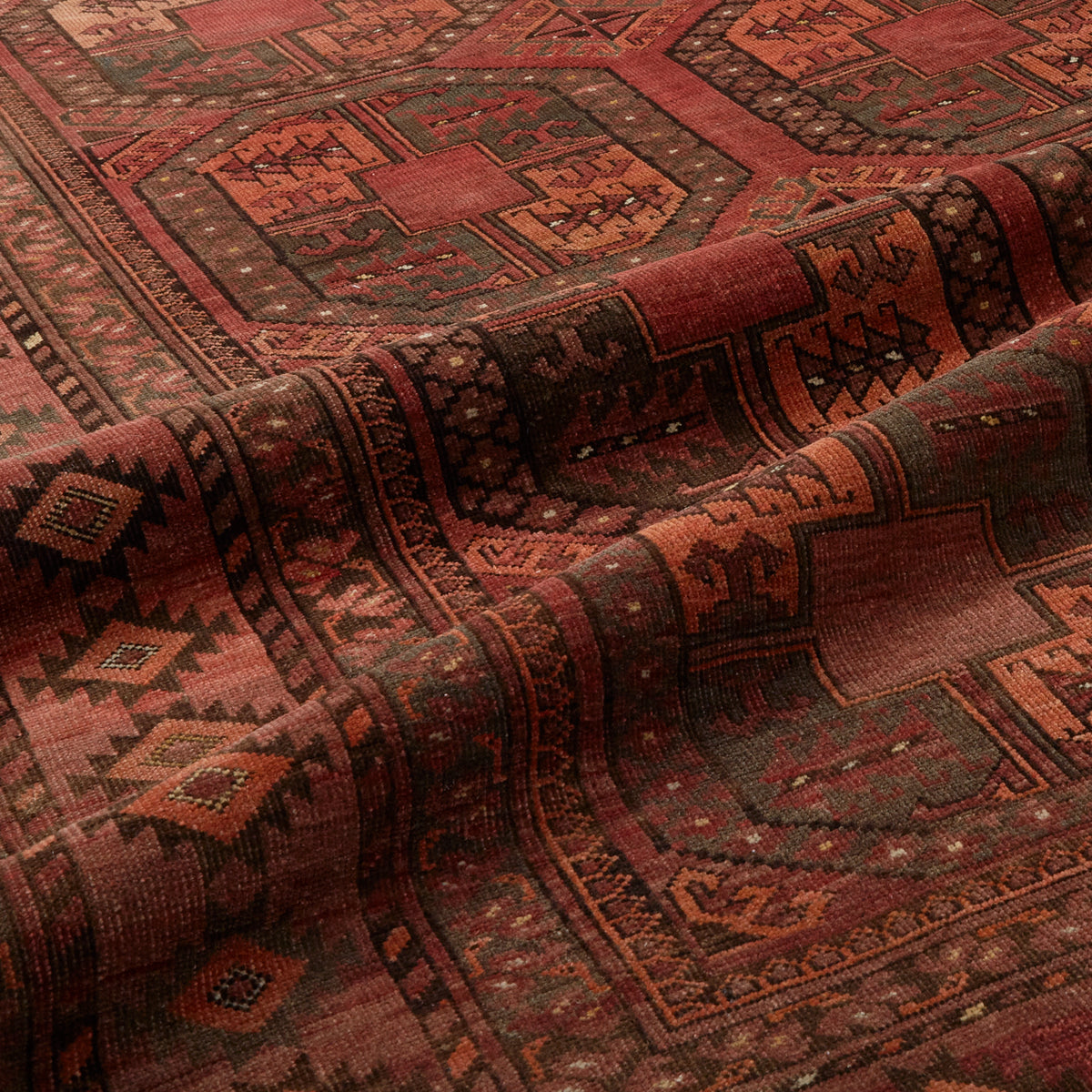 A close-up of a traditional patterned rug with geometric motifs in deep red, rust orange, brown, and muted green tones, shown with soft folds highlighting its texture.