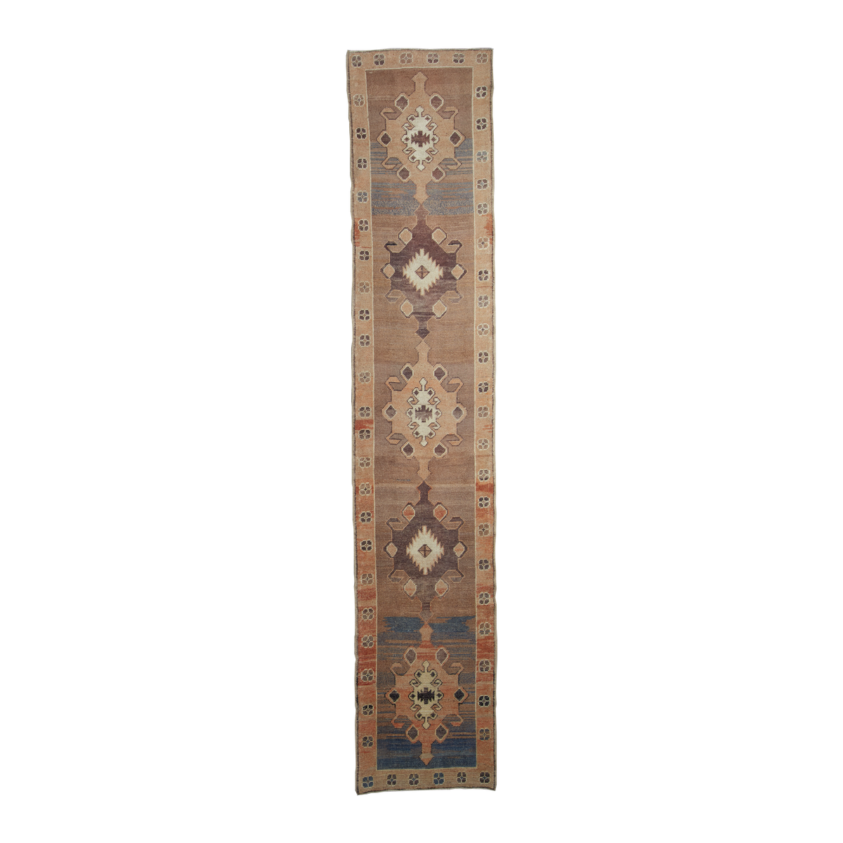 A long, narrow vintage-style runner rug with a beige base, featuring three large central diamond medallions in brown, blue, cream, and rust, bordered by geometric patterns on a white background.