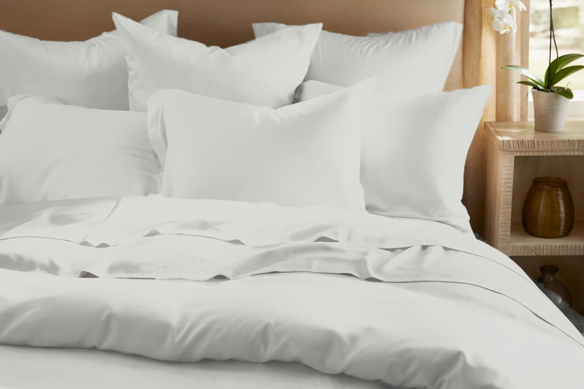 Legna Classic Bedding Set shown in Silver with a soft satin sheen, front view of a layered bed with euro and lumbar pillows, a flat sheet folded back over the duvet cover, and flanged pillowcases against a natural wood headboard.